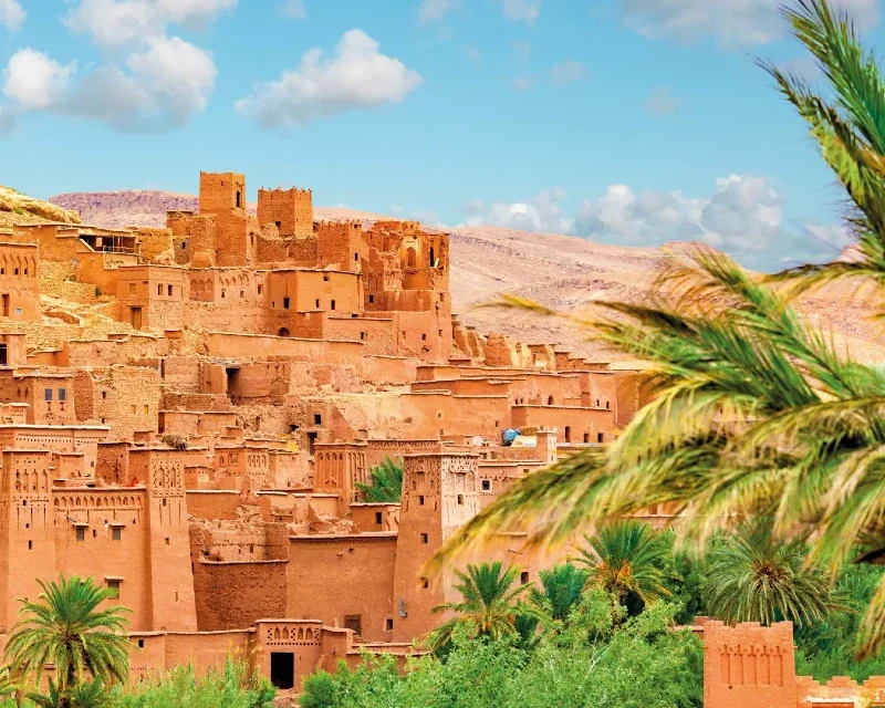 "The ancient clay city of Ait Benhaddou in Morocco, surrounded by lush palm trees under a bright blue sky."