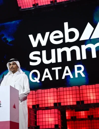 A speaker dressed in traditional Qatari attire delivering a keynote address at Qatar Web Summit 2025, with the event logo displayed prominently in the background.