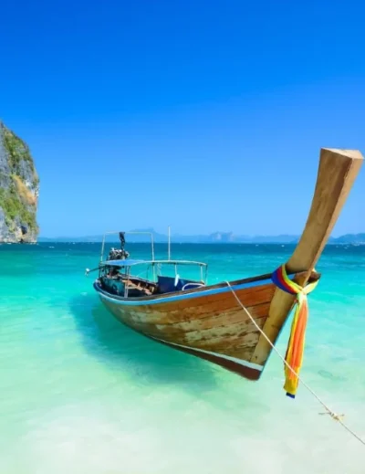 "A traditional Thai long-tail boat anchored in crystal-clear turquoise waters with a limestone karst island in the background, showcasing the beauty of Thailand's beaches."