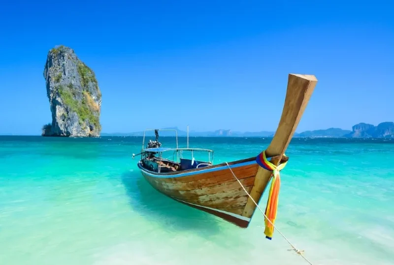 "A traditional Thai long-tail boat anchored in crystal-clear turquoise waters with a limestone karst island in the background, showcasing the beauty of Thailand's beaches."