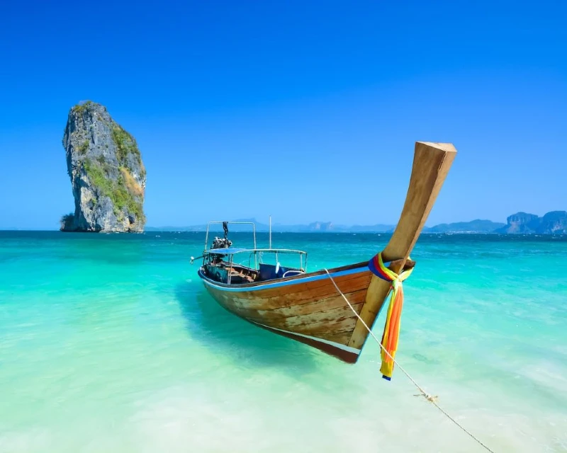 "A traditional Thai long-tail boat anchored in crystal-clear turquoise waters with a limestone karst island in the background, showcasing the beauty of Thailand's beaches."