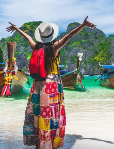 A woman in a colorful skirt and sunhat with arms raised, standing on a tropical beach in Thailand, facing traditional longtail boats anchored in turquoise water surrounded by limestone cliffs.