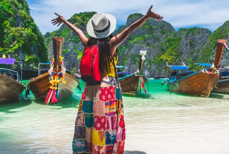 A woman in a colorful skirt and sunhat with arms raised, standing on a tropical beach in Thailand, facing traditional longtail boats anchored in turquoise water surrounded by limestone cliffs.