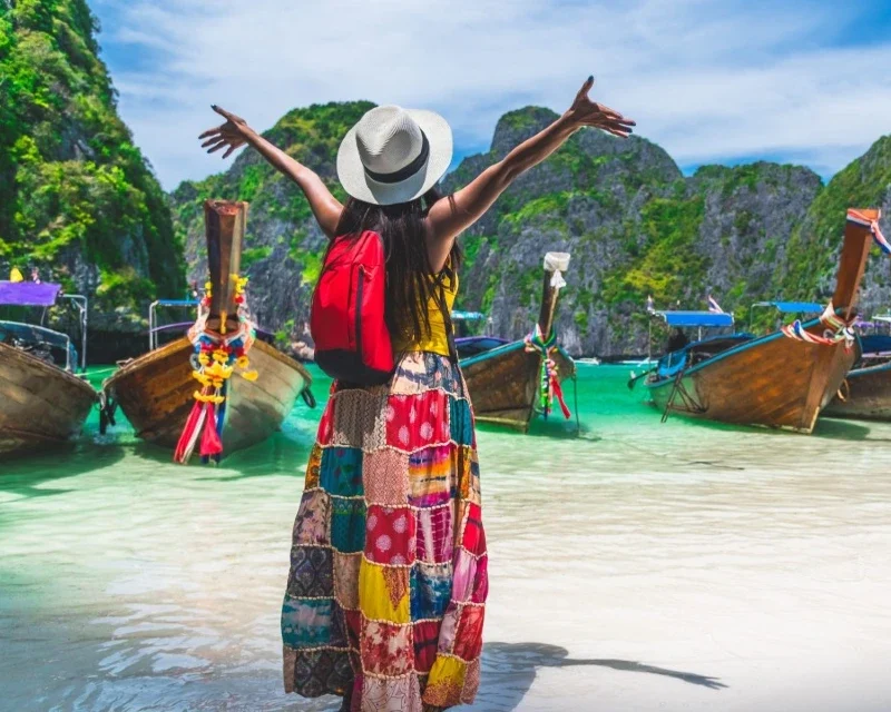 A woman in a colorful skirt and sunhat with arms raised, standing on a tropical beach in Thailand, facing traditional longtail boats anchored in turquoise water surrounded by limestone cliffs.