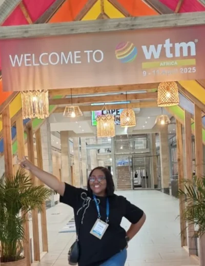 A smiling woman representing Lery Hago poses under the “Welcome to WTM Africa 2025” banner at the Cape Town International Convention Centre.