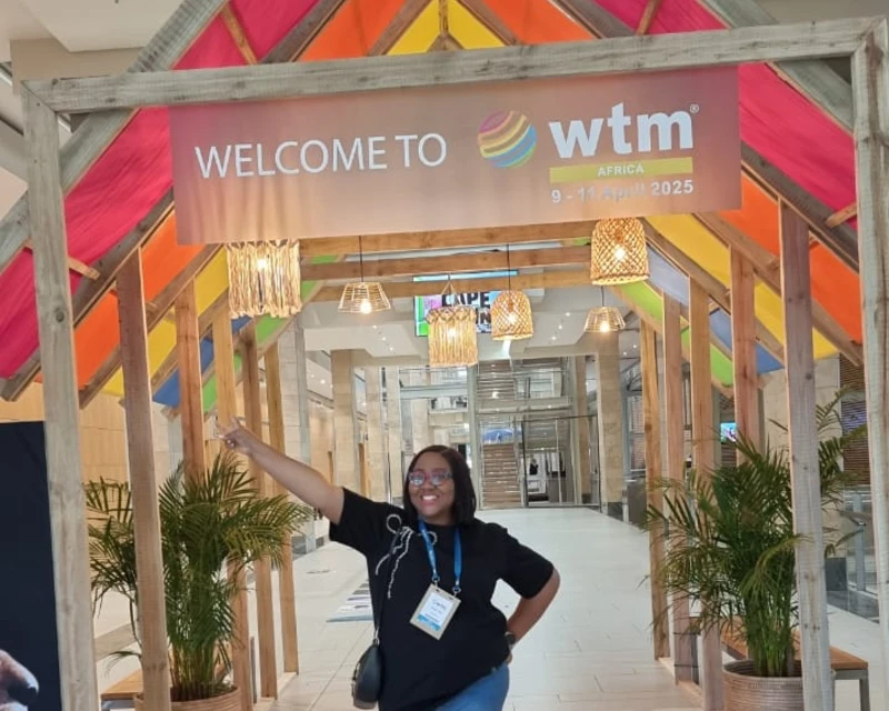 A smiling woman representing Lery Hago poses under the “Welcome to WTM Africa 2025” banner at the Cape Town International Convention Centre.