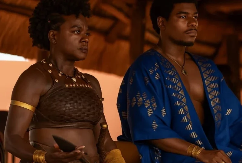 A powerful Dahomey warrior woman in traditional armor beside a regal man in blue royal attire, both seated in a historic African palace setting.