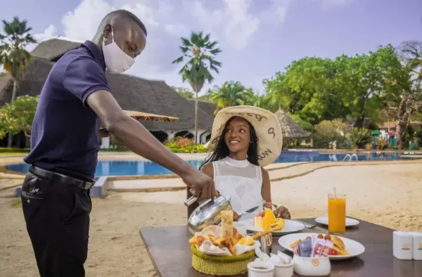 "Waiter in a mask serving a smiling young girl in a straw hat a tropical breakfast by a resort pool, with palm trees in the background."