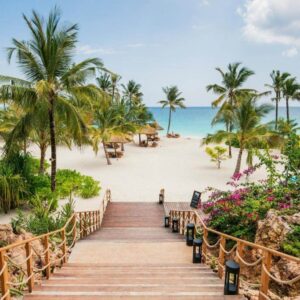 "Romantic Getaway Zanzibar: Wooden stairway leading down to a beautiful beach with white sand, palm trees, and turquoise waters, flanked by vibrant tropical flowers."