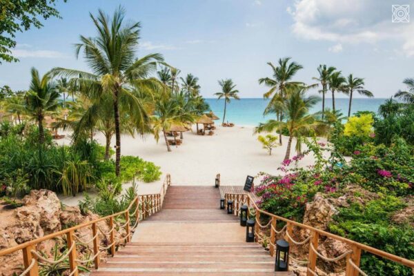 "Romantic Getaway Zanzibar: Wooden stairway leading down to a beautiful beach with white sand, palm trees, and turquoise waters, flanked by vibrant tropical flowers."