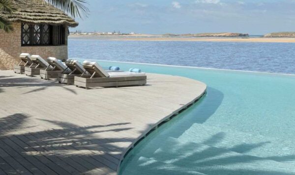 "Infinity pool merging with the sea horizon, featuring sun loungers on a wooden deck by a straw hut, embodying a Marrakech Love Getaway destination from www.leryhago.com."