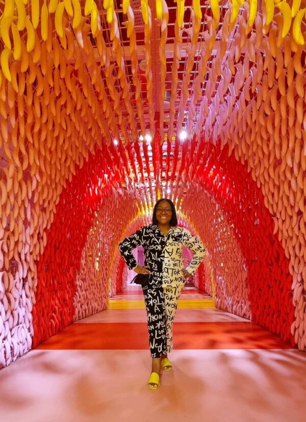 "A woman in a chic black-and-white outfit stands in the center of a vibrant corridor with cascading ribbons of yellow to red hues, part of the Christmas in Singapore Package."
