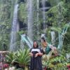 "A cheerful woman in a stylish outfit stands before a majestic waterfall surrounded by lush greenery, part of the Christmas in Singapore Package."