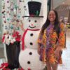 "A delighted woman in a colorful dress poses next to a cheerful snowman decoration amid a Christmas display in Singapore."