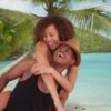 "A couple laughing joyfully as the man carries the woman on his back on a tropical beach, palm trees and turquoise water in the background."