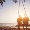 "Silhouette of a couple sitting on a beach swing at sunset, with the ocean and palm leaves in the foreground."