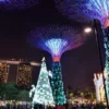 "Spectacular view of Singapore's Gardens by the Bay with towering Supertrees adorned in Christmas lights, with a large lit Christmas tree as the centerpiece."