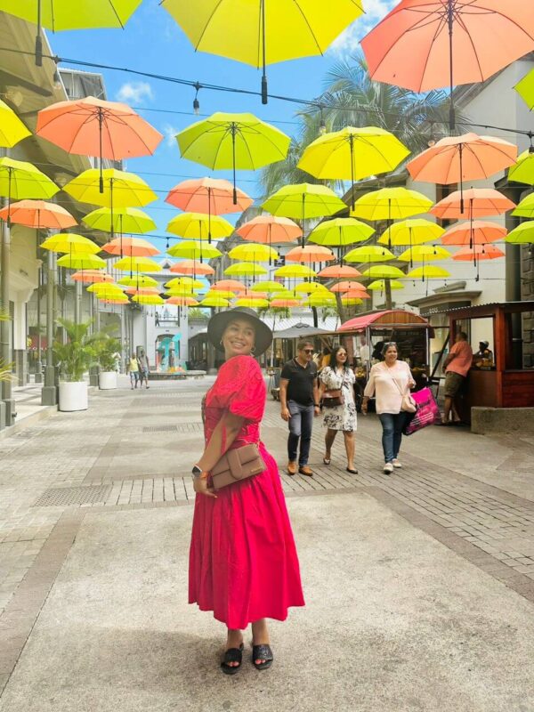"Woman in a vibrant red dress under a colorful canopy of umbrellas in a street of Nairobi Mauritius, encapsulating the festive spirit of a Pre-Christmas Escape."