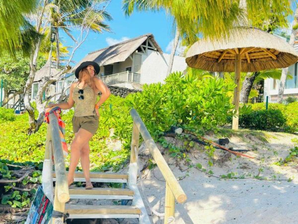 "A stylish traveler poses on wooden stairs leading to a serene beach resort in Nairobi Mauritius, surrounded by lush greenery and a thatched umbrella, epitomizing a Pre-Christmas Escape."