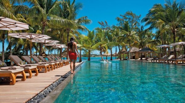"A woman strolls by a luxurious infinity pool surrounded by palm trees and sun loungers in an idyllic tropical resort setting."