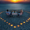 "A couple enjoys a romantic sunset dinner on the beach, encircled by a heart-shaped array of glowing candles on the sand."