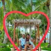 "A couple smiles within a large heart-shaped frame on a Maldivian beach, surrounded by lush palms, encapsulating the romance of their villa stay. Your perfect getaway at www.leryhago.com."