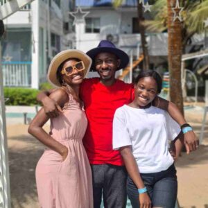 "A joyful trio poses under the tropical sun, with a woman in a chic hat and sundress, a man in a vibrant red shirt and hat, and a young lady in a casual white tee and shorts, at a Nigerian Romantic Resort, with palm trees and a beach house in the background."