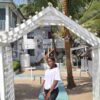 "A confident young woman in a white T-shirt and black shorts stands poised at the entrance of a Nigerian Romantic Resort, with elegant palm trees and a charming white beach house in the background."