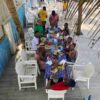 "A group of guests enjoy a communal feast at a long table set with an array of delicious dishes under the shade of palm trees at a Nigerian Romantic Resort."