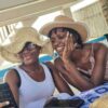 "Two women share a candid moment, one in a stylish straw hat and the other in a chic pink dress, both enjoying a sunny day at a Nigerian Romantic Resort."