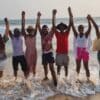 "A jubilant group of friends with raised hands stands in the surf, celebrating their bond at the shoreline of a Nigerian Romantic Resort."