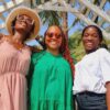 "Three women in stylish summer attire, with radiant smiles and sunglasses, pose under a white lattice archway amidst palm trees at a Nigerian Romantic Resort."