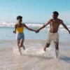 "Joyful African-American couple holding hands and running through the sea on a sunny day, an idyllic scene from a Romantic Turkey Tour at www.leryhago.com." Caption: "Find joy in the simplicity of love with the Romantic Turkey Tour by www.leryhago.com – where every step is a dance on the shores of Turkey."