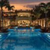 "A luxurious resort pool illuminated by ambient lights, flanked by palm trees and a tropical villa at dusk."