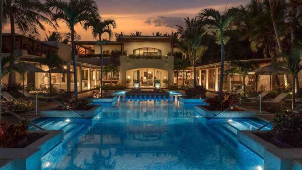 "A luxurious resort pool illuminated by ambient lights, flanked by palm trees and a tropical villa at dusk."