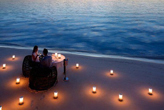 "A couple enjoys a romantic candlelit dinner on the beach, with candles in the sand and the ocean's gentle waves in the twilight."