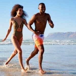 "A cheerful couple running along the shore, their feet splashing in the shallow waves, mountains in the distance."