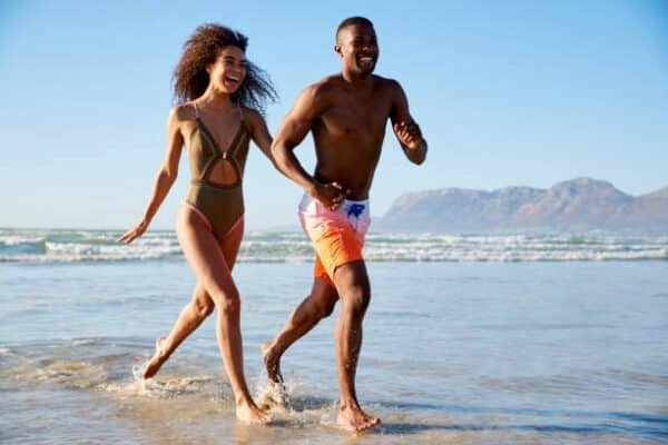 "A cheerful couple running along the shore, their feet splashing in the shallow waves, mountains in the distance."
