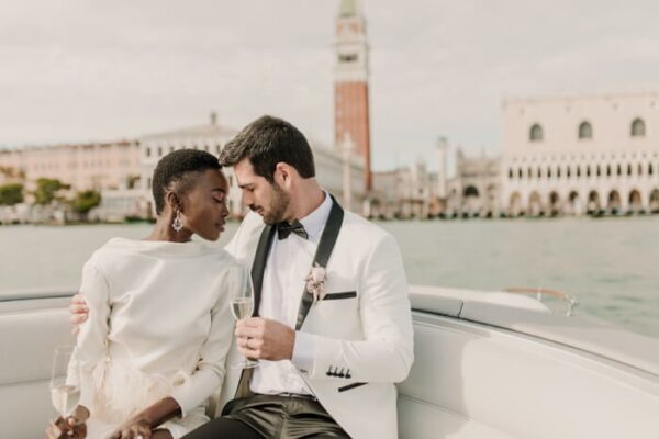 "Elegant couple sharing a toast on a boat with Venice's iconic landmarks in the background, epitomizing a sophisticated Venice Valentine's Getaway - plan at www.leryhago.com."