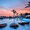 "Sunset hues painting the sky above a luxurious resort pool lined with palm trees, offered by Paris Love Getaway on www.leryhago.com."