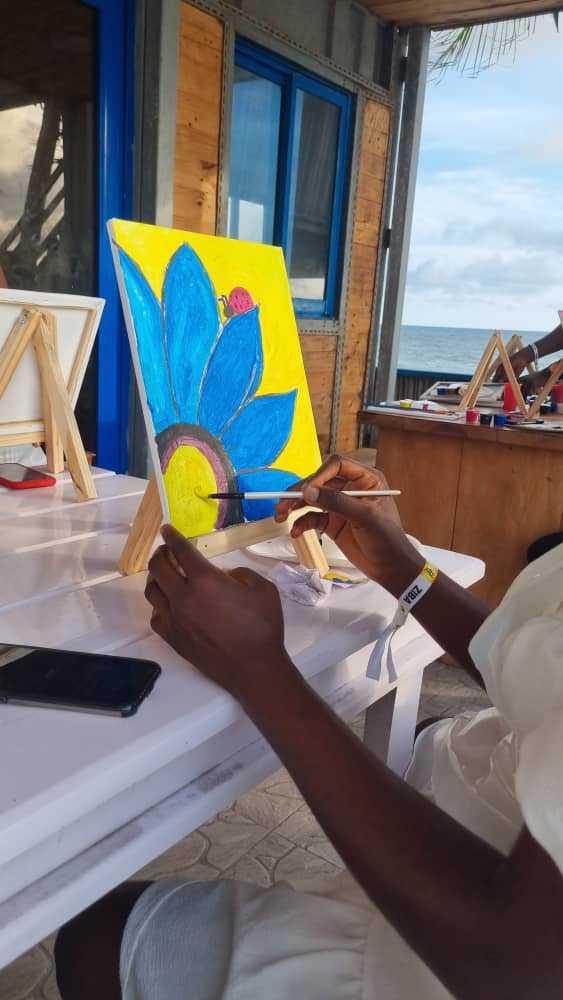 "Artist painting a vibrant blue flower on a canvas at a beachside workshop, embodying the spirit of a Naija Easter Getaway, showcased on www.leryhago.com."