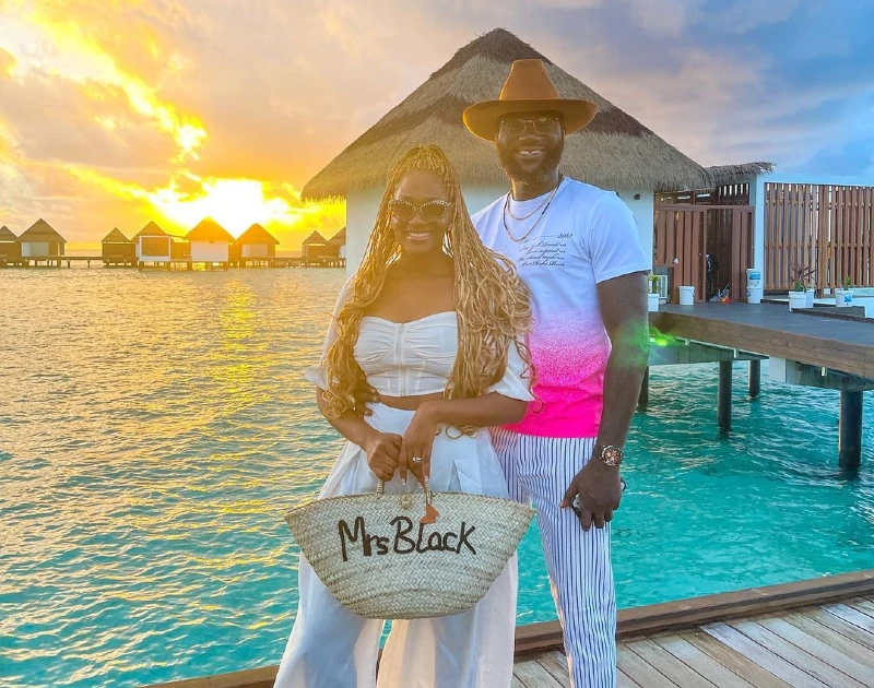 A smiling couple posing at sunset by the turquoise waters of a luxurious overwater bungalow, holding a customized bag that says 'Mrs. Black.