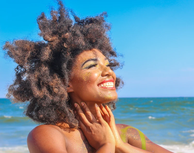 A woman with natural curly hair smiling under the bright sun on a beach with the ocean in the background.