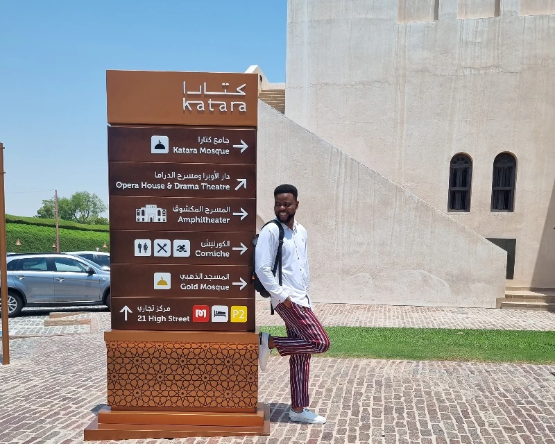 Man standing next to a signpost in Katara Cultural Village, Doha, highlighting tourist attractions with Lery Hago Travels