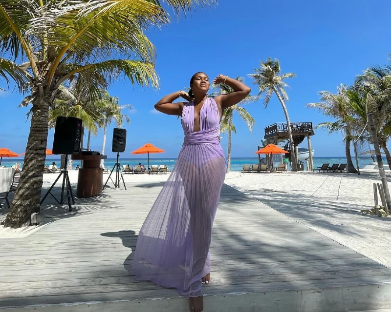 A woman in a flowing lavender dress enjoying the sunshine at a tropical all-inclusive beach resort, surrounded by palm trees and beach umbrellas.