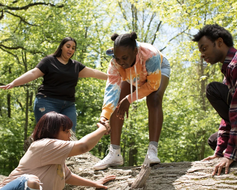 "A group of people helping each other climb over a log in a forest during a family vacation, showcasing teamwork and adventure. Your essential family vacation packing list: what to bring when travelling with kids of all ages - Lery Hago Travels."