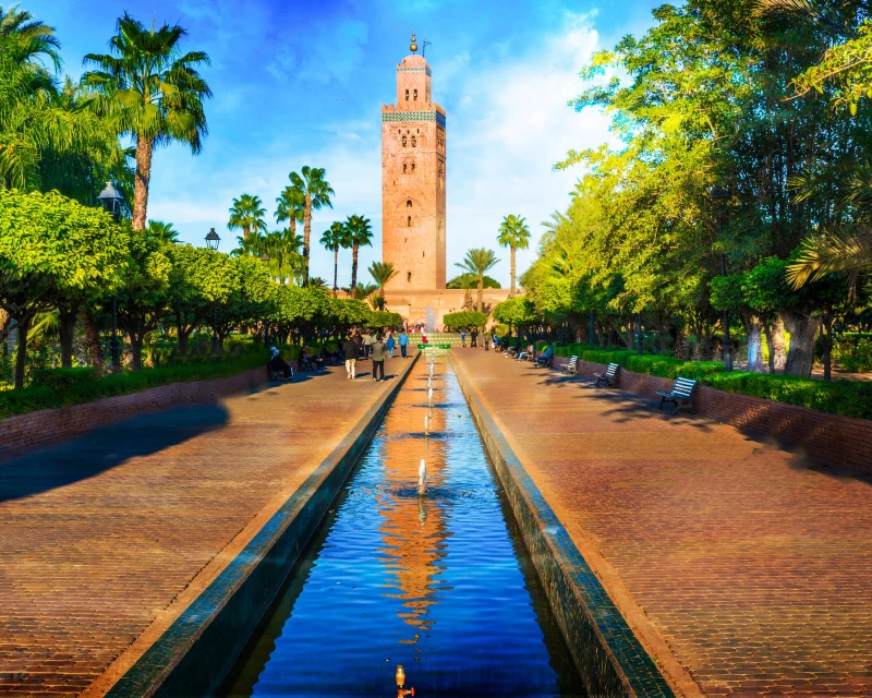 Scenic view of the Koutoubia Mosque in Marrakech, Morocco, with a tranquil water feature and lush gardens, captured on a sunny day.