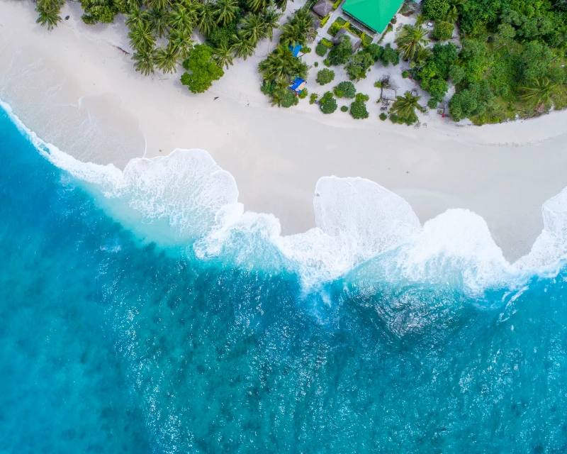 "A mesmerizing aerial view of waves crashing onto a Maldives beach, bordered by lush greenery and a secluded resort, perfect for first-timers with Lery Hago Travels."