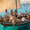 An elevated, cinematic drone shot captures a traditional wooden dhow boat sailing through the crystal-clear, vibrant turquoise waters of Zanzibar. Onboard, a group of stylish African men and women in their late 20s and 30s celebrate under a warm golden hour sun. They are dressed in elegant resort wear, including white linen shirts, patterned sarongs, and summer hats. One man sits prominently at the bow, joyfully waving toward the camera, while others lounge on the deck, laughing and enjoying the breeze. The large, weathered sail is filled with wind, and the surrounding ocean transitions from a glowing electric teal to a deep sapphire, creating a luxurious and aspirational travel aesthetic.